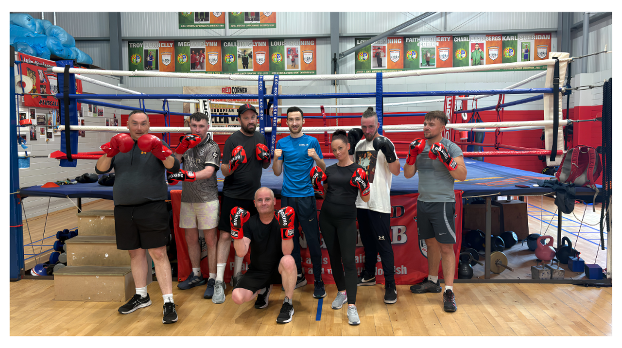 A group of eight people in boxing gloves poses in front of a boxing ring in a gym.